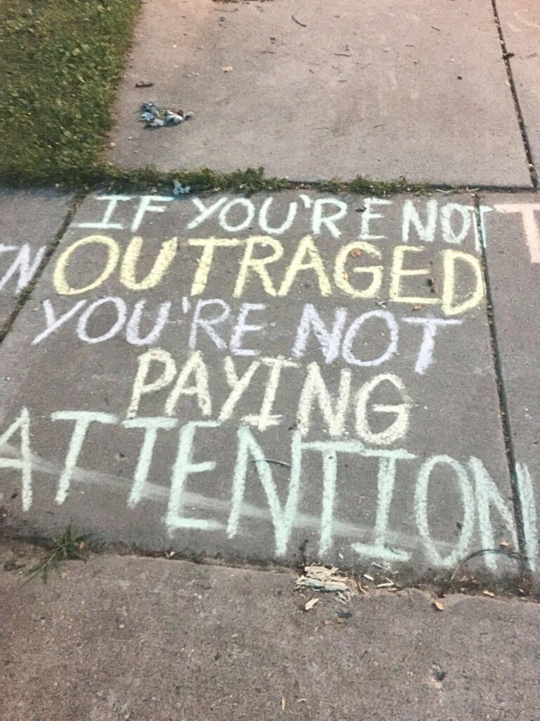 Sidewalk chalk writing "If you are not outraged, you are not paying attention."