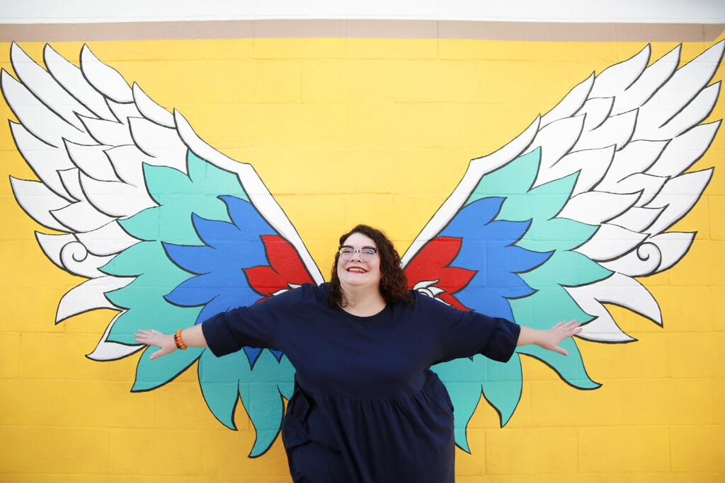 Me standing arms wide in front of a wall mural of brightly colored wings